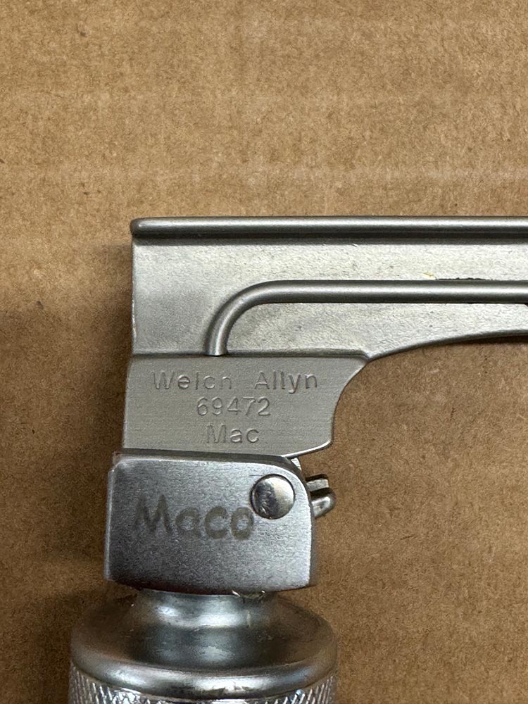Close-up of a metal lighter head with engraved text: 'Welch Allyn 69472 Mac' on a silver latch, against a brown background.
