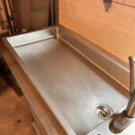 Large stainless steel utility sink in a garage/workshop with a high faucet and round drain drain visible in the basin edge.