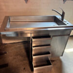 Stainless steel utility sink with a tall curved faucet and a five-drawer metal cabinet beneath.