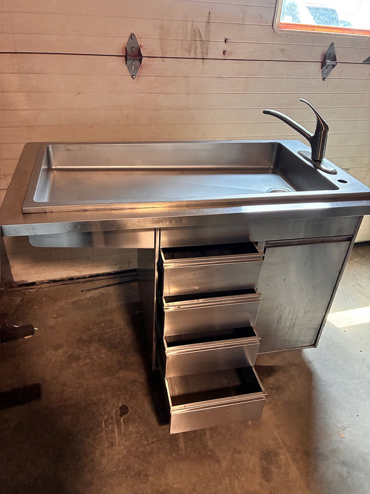 Stainless steel utility sink with a tall curved faucet and a five-drawer metal cabinet beneath.
